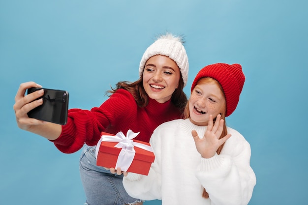 Menina bonita jovem com sardas na camisa branca e boné vermelho, acenando com a mão, abraçando o presente e tira uma selfie com a irmã sorridente