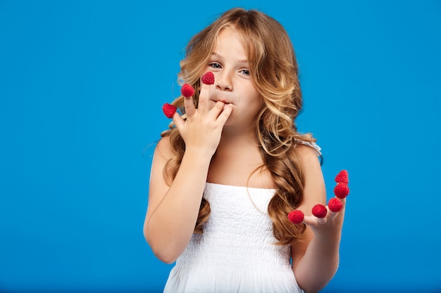 Menina bonita jovem comendo framboesa sobre parede azul