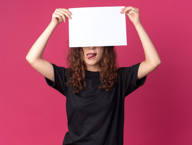 Menina bonita jovem e brincalhona levantando um papel em branco segurando-o na frente dos olhos, mostrando a língua isolada na parede carmesim