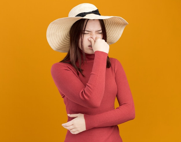 Menina bonita jovem enojada com chapéu de praia fazendo gesto de mau cheiro com os olhos fechados, isolada na parede laranja com espaço de cópia