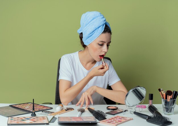 Menina bonita jovem sentada à mesa de maquiagem com ferramentas de maquiagem e com a toalha na cabeça, olhando para o espelho, passando batom vermelho e tocando mesa no espaço verde oliva