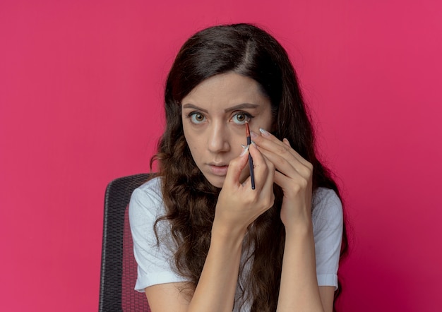 Menina bonita jovem sentada à mesa de maquiagem com ferramentas de maquiagem, tocando o rosto e aplicando a sombra com pincel de maquiagem, olhando para a câmera isolada no fundo carmesim