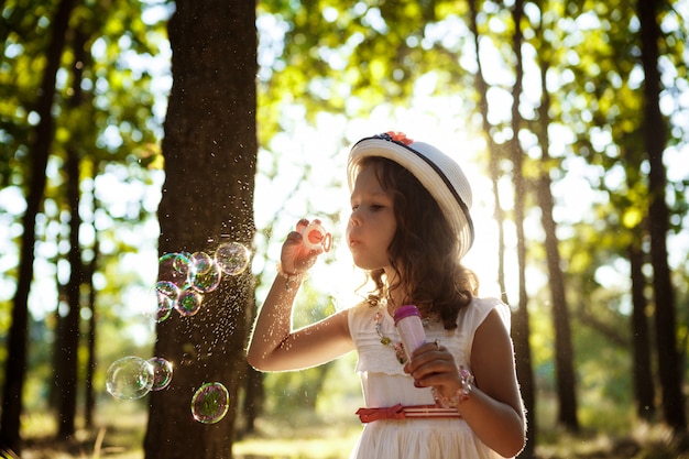 Menina bonita jovem soprando bolhas, andando no parque ao pôr do sol.