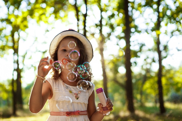 Menina bonita jovem soprando bolhas, andando no parque ao pôr do sol.
