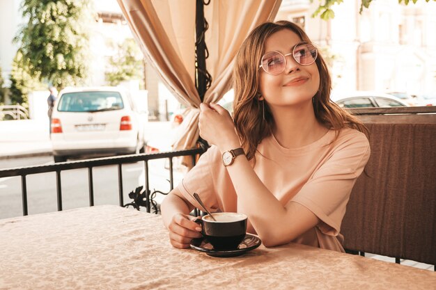 Menina bonita jovem sorridente hipster em roupas da moda no verão. Mulher despreocupada, sentado no terraço varanda café e bebendo café. Modelo positivo, se divertindo e sonhos