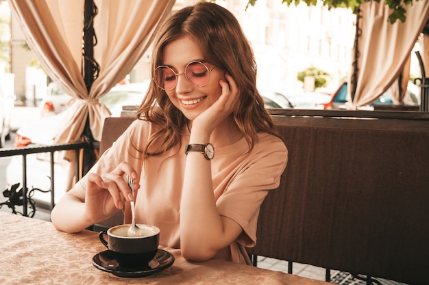 Menina bonita jovem sorridente hipster em roupas da moda no verão. Mulher despreocupada, sentado no terraço varanda café e bebendo café. Modelo positivo, se divertindo e sonhos