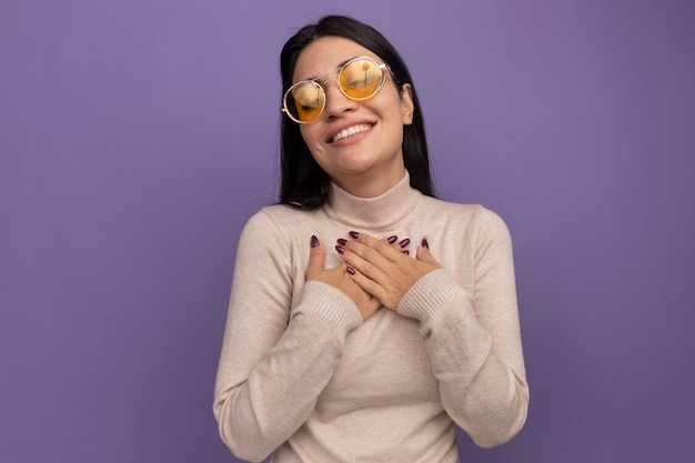 Foto grátis menina bonita morena caucasiana satisfeita com óculos de sol colocando as mãos no peito roxo