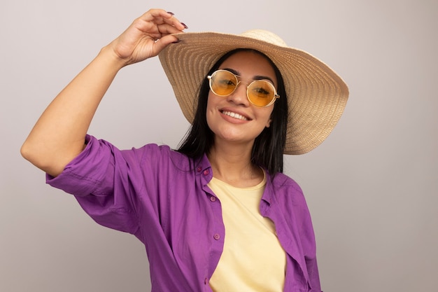 Menina bonita morena caucasiana sorridente com óculos de sol e chapéu de praia coloca a mão no chapéu branco