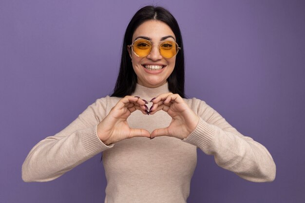 Menina bonita morena caucasiana sorridente usando óculos de sol, gesticulando coração mão assinar roxo