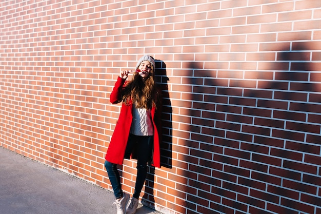 Foto grátis menina bonita morena de comprimento total com casaco vermelho na luz do sol na parede do lado de fora. ela usa chapéu de tricô, lambe os lábios vermelhos de pirulito, mantém os olhos fechados.