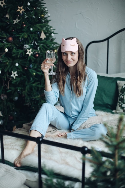 Menina bonita morena de pijama e máscara de dormir, levantando a taça de champanhe, sentada na cama. xmas.