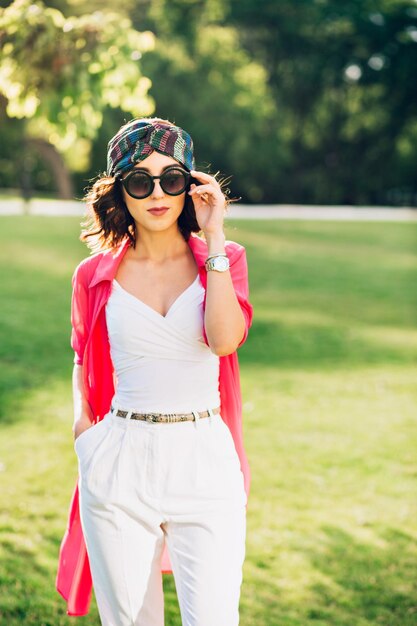 Menina bonita morena em bandana está posando no parque de verão. Ela usa roupas brancas, óculos escuros e casaco comprido rosa. Ela segura os óculos de sol no rosto e mantém a mão no bolso.