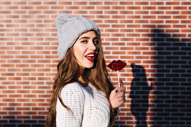 Foto grátis menina bonita morena retrato de suéter branco e chapéu de malha na parede do lado de fora. ela segura os lábios vermelhos de pirulito, sorrindo.