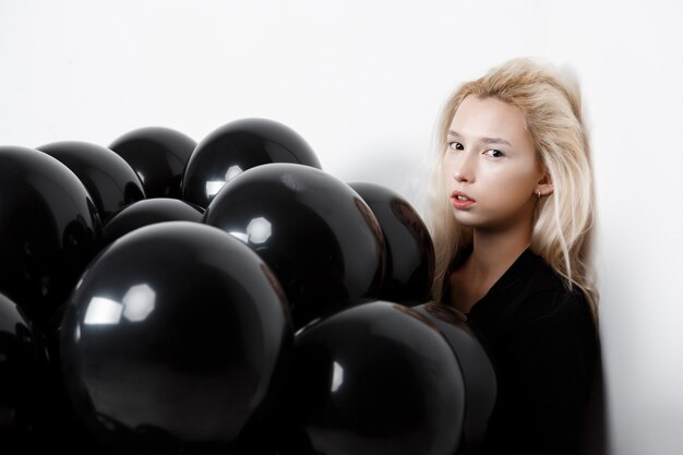 Menina bonita nova que senta-se em balões pretos sobre a parede branca.