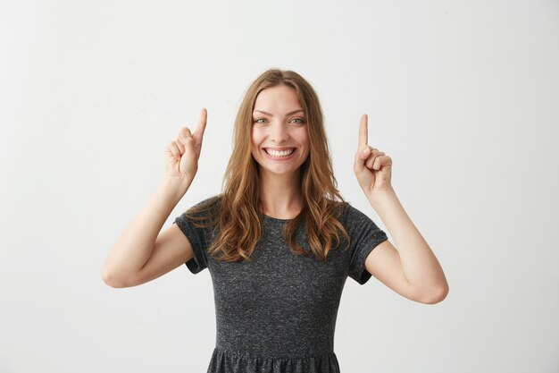 Menina bonita positiva nova que sorri apontando os dedos acima.