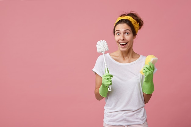 Menina bonita se sentindo chocada com notícias inesperadas enquanto faz a limpeza geral, usando luvas de borracha, lavando o banheiro, segurando escova e esponja, olhando, mantendo a boca bem aberta