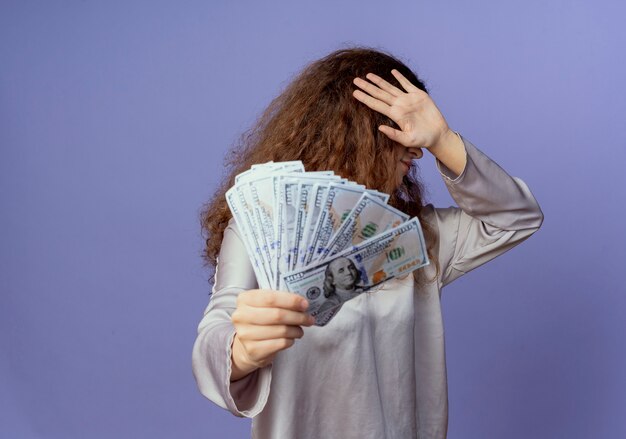 Menina bonita segurando dinheiro e rosto coberto com a mão isolada em azul
