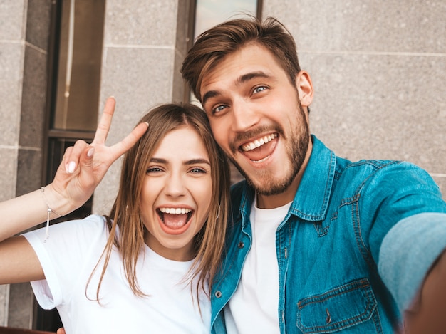 Menina bonita sorridente e seu namorado bonito em roupas de verão casual. família feliz, tendo selfie auto-retrato de si na câmera do smartphone. mostra sinal de paz e piscando na rua