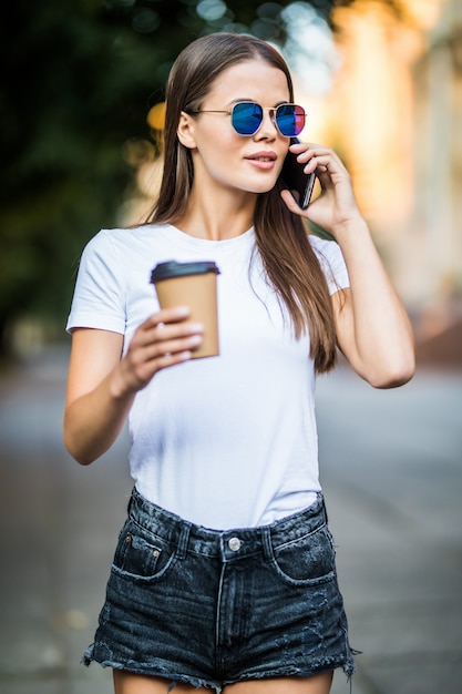 Menina bonita sorridente, falando no telefone celular e tomando café na rua