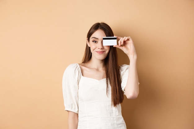Menina bonita sorridente mostrando cartão de crédito de plástico sobre o olho e parecendo satisfeito em pé no bac bege.