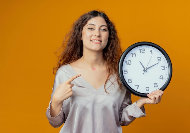 Menina bonita sorridente segurando e apontando para o relógio de parede isolado na parede amarela