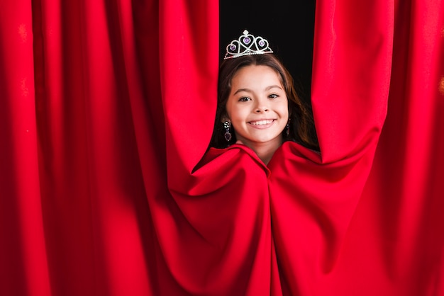 Menina bonita sorridente usando coroa espreitar da cortina vermelha