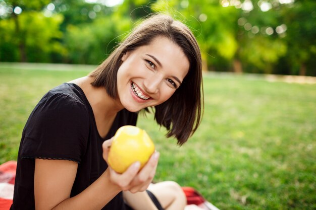 Menina bonita sorrindo, segurando a maçã no piquenique no parque.