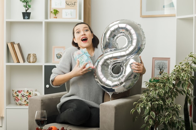Foto grátis menina bonita surpresa no dia da mulher feliz segurando o balão número oito com um presente sentado na poltrona na sala de estar
