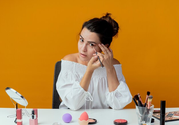 Menina bonita surpresa sentada à mesa com ferramentas de maquiagem, aplicando sombra com pincel de maquiagem, parecendo isolada na parede laranja