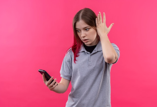 Menina bonita surpresa vestindo camiseta cinza olhando para o telefone na mão em fundo rosa isolado