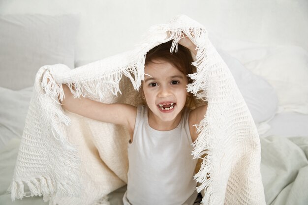 Menina bonitinha brincando na cama com um cobertor.