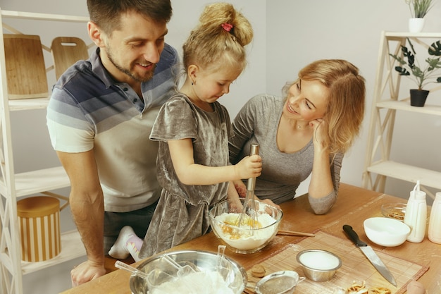 Menina bonitinha e seus lindos pais preparando a massa para o bolo na cozinha em casa. Conceito de estilo de vida familiar