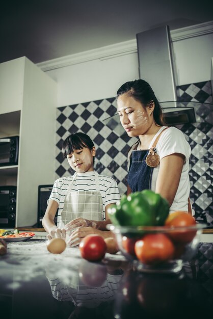 Menina bonitinha e sua mãe em aventais aplainar a massa usando um rolo na cozinha