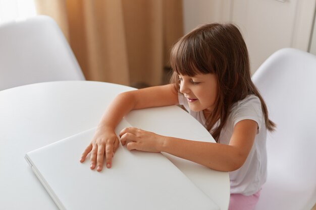 Menina bonitinha está sentada à mesa com seu laptop branco fechado, criança do sexo feminino de cabelos escuros, posando na sala de luz, usando o fechamento de estilo casual, tiro interno.