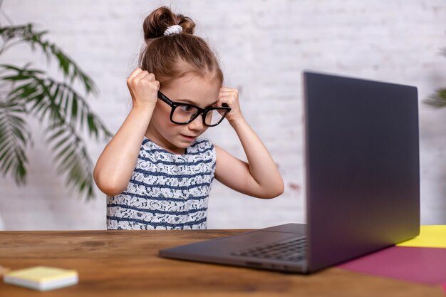 Menina bonitinha está sentada à mesa com seu laptop preto e usando óculos em casa. Uma menina criança fazendo lição de casa ou jogando