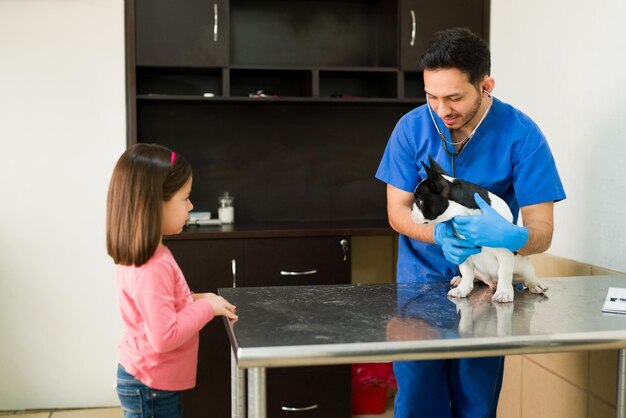 Menina bonitinha fazendo perguntas sobre seu boston terrier doente para um veterinário masculino profissional. Veterinário de homem hispânico examinando um cachorro pequeno na clínica de animais