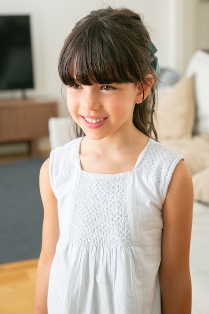 Menina bonitinha no vestido branco de pé e sorrindo na sala de estar.