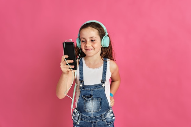 Menina bonitinha posando com um telefone e fones de ouvido em uma parede rosa