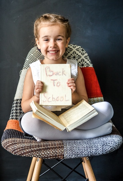 Menina bonitinha sentada em uma linda cadeira com um livro nas mãos, o conceito de educação e vida escolar