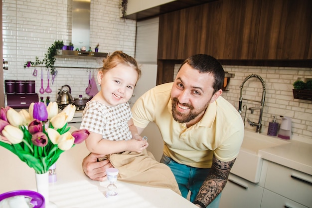 Menina bonitinha sentada na mesa da cozinha com seu pai sorridente feliz olhando para a câmera sorrindo alegremente na cozinha em casa se divertindo juntos conceito de família feliz