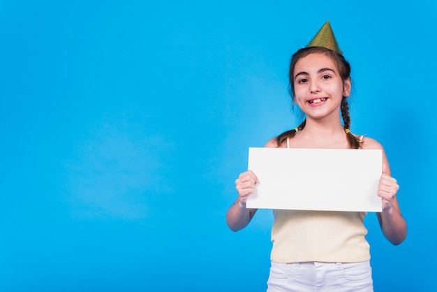 Menina bonito de sorriso que desgasta o chapéu do partido que prende o cartão em branco na mão na frente do papel de parede colorido