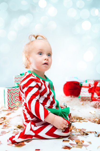 Menina bonito perto de chapéu de Papai Noel posando sobre fundo de Natal com decoração. Sentado no chão com uma bola de Natal.