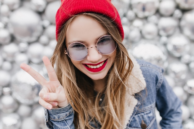 Menina branca interessada com sorriso alegre, posando com o símbolo da paz. Foto de close-up de uma senhora atraente em jaqueta jeans, brincando durante a sessão de fotos na parede brilhante.