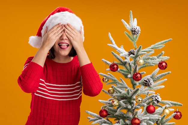 Foto grátis menina brincalhona com chapéu de papai noel em pé perto da árvore de natal decorada, cobrindo os olhos com as mãos, mostrando a língua isolada na parede laranja