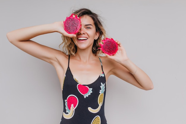 Menina bronzeada feliz com tatuagem de braço segurando pitaya vermelha Foto interna de mulher loira despreocupada sorrindo durante sessão de fotos com fruta do dragão