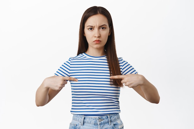 Menina carrancuda chateada apontando os dedos para o logotipo no centro de sua camisa, amuada decepcionada, não gosta de smth, expressa antipatia e desacordo, de pé sobre fundo branco