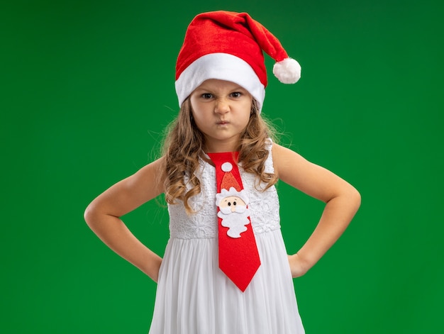 Foto grátis menina carrancuda usando chapéu de natal com gravata colocando as mãos no quadril isolado sobre fundo verde