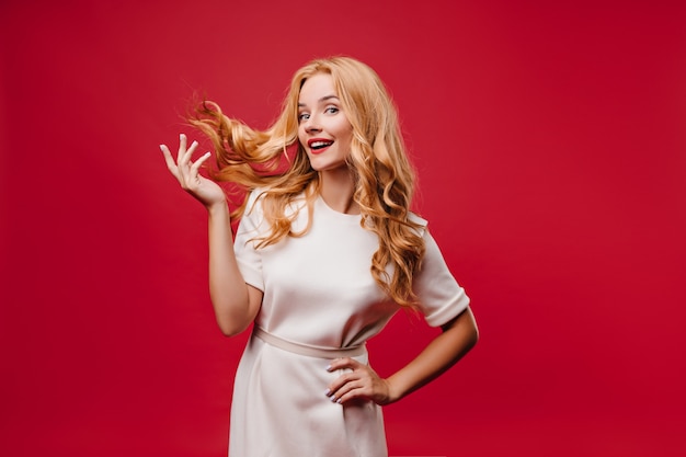 Foto grátis menina caucasiana atraente brincando com seus longos cabelos na parede vermelha. foto interna de uma mulher atraente e elegante no vestido.