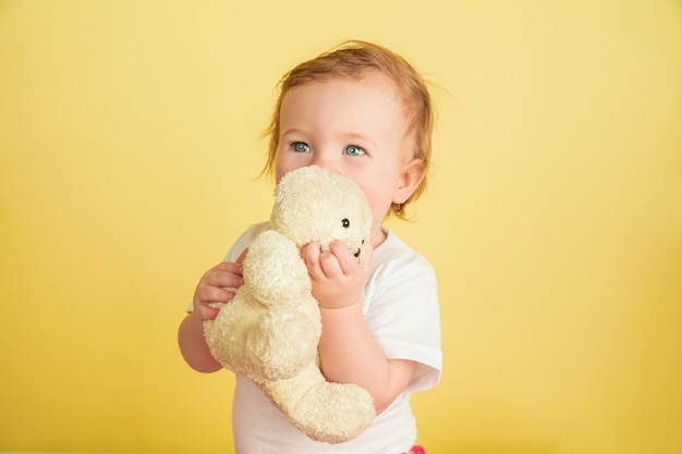 Menina caucasiana, crianças isoladas em fundo amarelo do estúdio. Retrato de criança fofa e adorável, bebê brincando com ursinho de pelúcia.