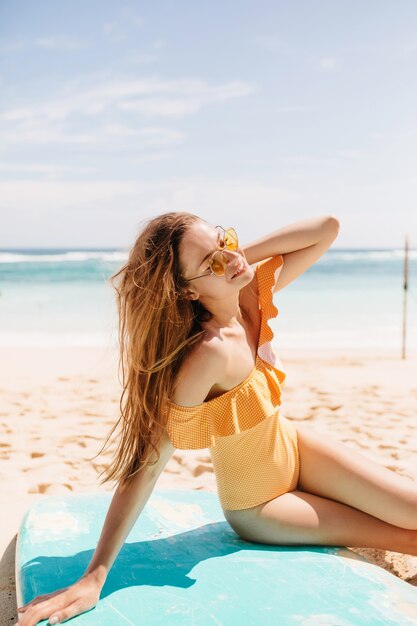 Menina caucasiana de cabelos castanhos positiva posando na praia com um sorriso feliz. Retrato ao ar livre da incrível mulher morena em trajes de banho laranja relaxantes depois de surfar no oceano.